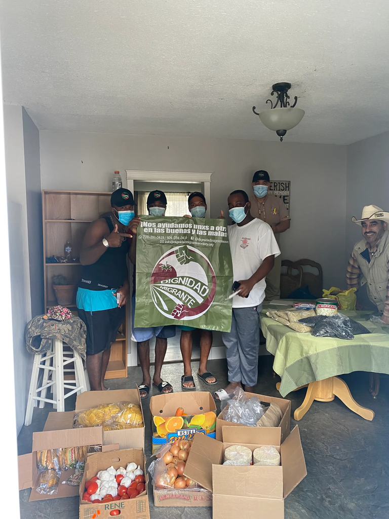 6 people stand in front of boxes of food. they are wearing medical masks. they are holding a dignidad migante sign.